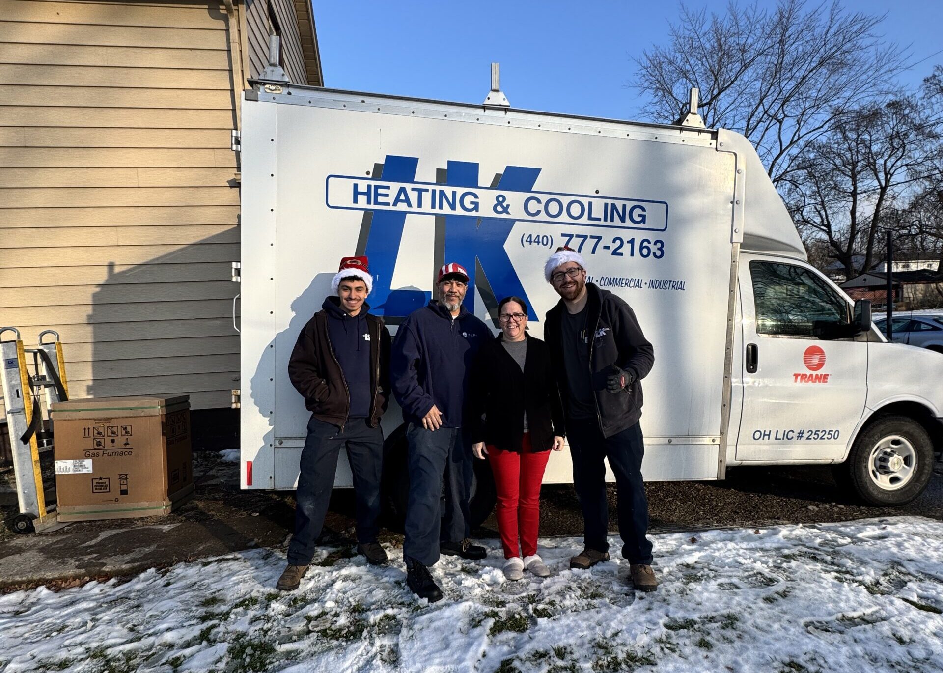 two install technicians wearing Santa hats, standing with the customers who won the free furnace giveaway, a man and woman. Behind them is the LK Heating and Cooling Truck. There's snow on the ground.